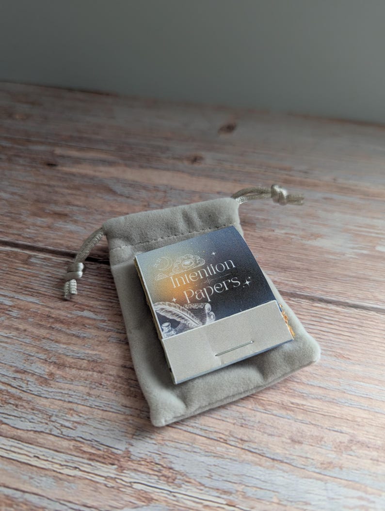 May include: A small, gray velvet pouch with a drawstring closure holds a booklet labeled "Intention Papers." The booklet has a starry night sky design and a feather illustration. The items rest on a wooden surface.
