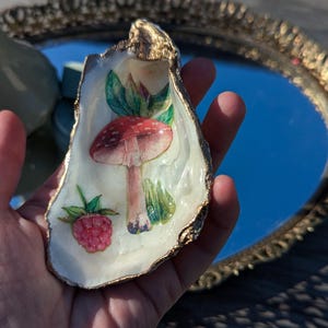 Puede incluir: Una concha de ostra blanca con un borde dorado, pintada con un hongo rojo y blanco y una frambuesa verde. La concha es un pequeño plato para baratijas.