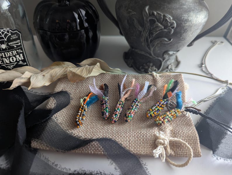 May include: Five beaded corn pendants with colourful beads and ribbon accents rest on a burlap surface. The pendants are in various colours, including blue, orange, and green. A black ribbon and a decorative pumpkin are in the background.