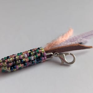 May include: A beaded corn charm with a silver clasp and leather and fabric accents. The corn is made of colourful beads in shades of green, pink, purple, and yellow. The charm has a brown leather piece with peach and lavender fabric.
