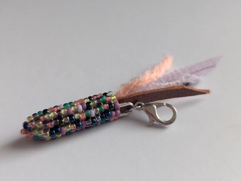 May include: A beaded corn charm with a silver clasp and leather and fabric accents. The corn is made of colourful beads in shades of green, pink, purple, and yellow. The charm has a brown leather piece with peach and lavender fabric.