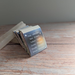 May include: A stack of small, rectangular cards with text and star graphics. The cards are labeled with colors and words like "Affection," "Protect," and "Manifest." The cards are on a wooden surface.