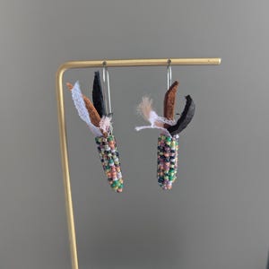 May include: Handmade beaded earrings, each resembling a small corn cob. The earrings are crafted with a variety of colourful beads, including green, purple, orange, and white. They also feature fabric leaves in black, brown, and white.