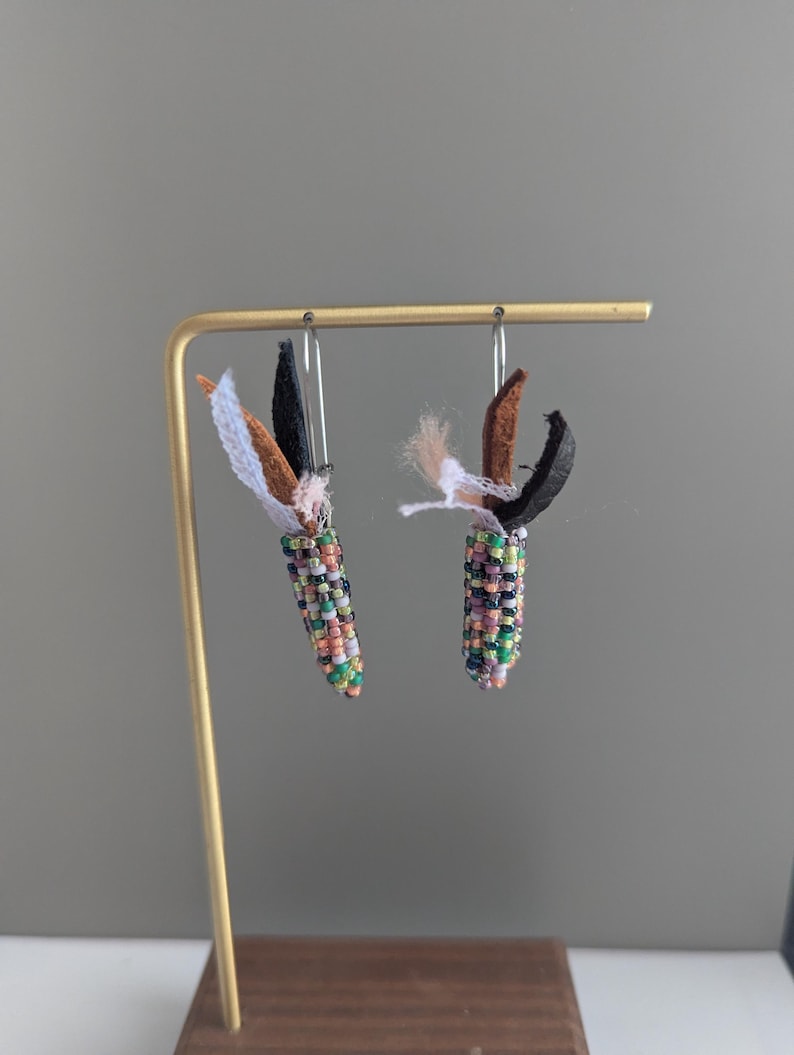 May include: Handmade beaded earrings, each resembling a small corn cob. The earrings are crafted with a variety of colourful beads, including green, purple, orange, and white. They also feature fabric leaves in black, brown, and white.