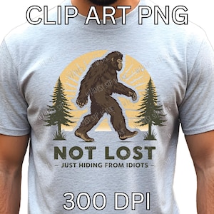 May include: A light gray t-shirt featuring a Bigfoot graphic walking in front of a yellow sun and two green pine trees. The text "NOT LOST - JUST HIDING FROM IDIOTS" is below the graphic. The words "CLIP ART PNG" and "300 DPI" are at the top and bottom.