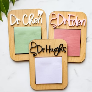 May include: Three wooden memo boards with rectangular frames. Each board has a different name in cursive lettering: "Dr. Chen", "Dr. Eden", and "Dr. Hughs". The "Dr. Chen" board has a tooth icon, and the "Dr. Eden" board has a stethoscope icon. Each board has a colored sticky note.