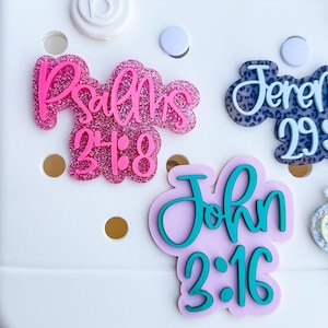May include: Close-up of decorative bag charms with religious scripture. The charms feature the text "Psalms 24:8", "Jeremiah 29:11", and "John 3:16" in various colors and fonts. The charms are attached to a white bag.