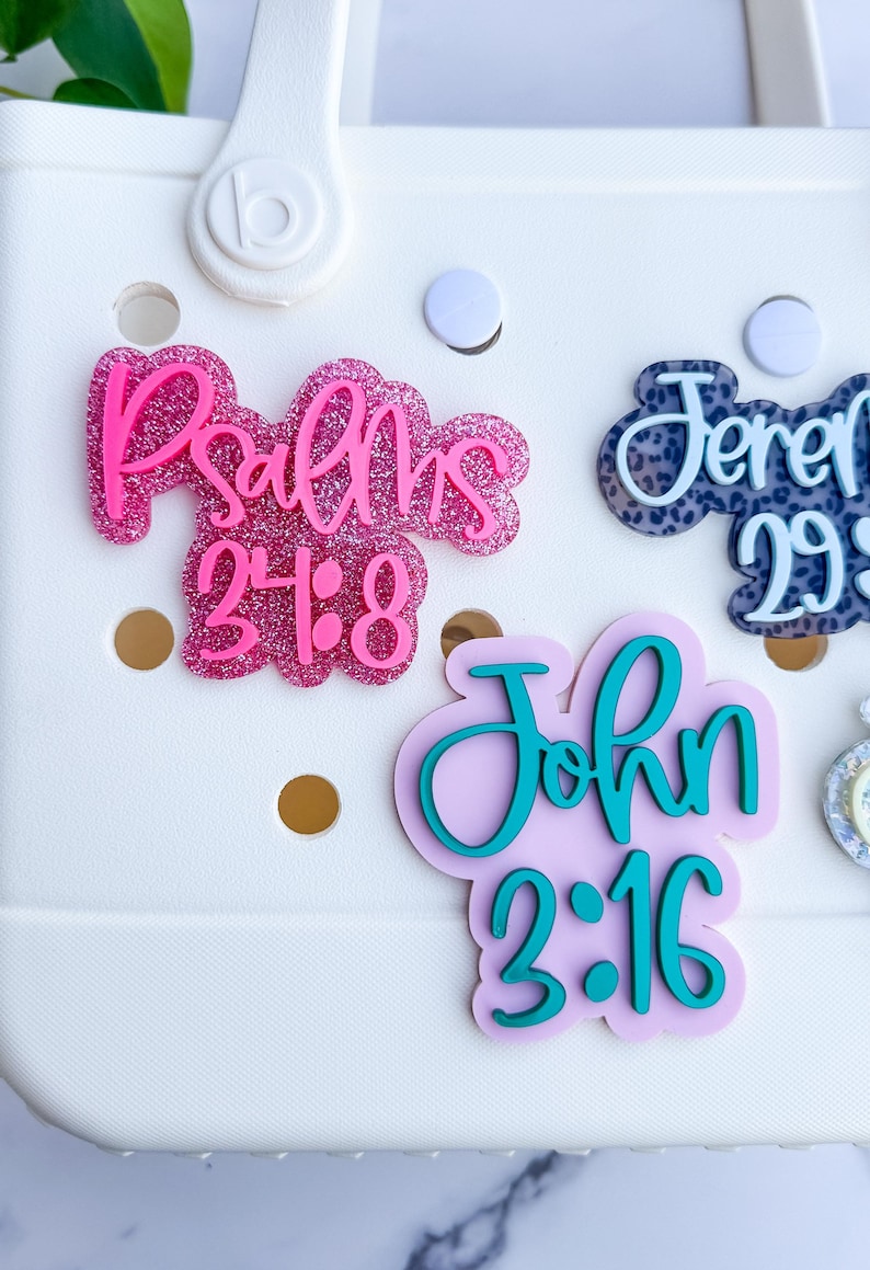 May include: Close-up of decorative bag charms with religious scripture. The charms feature the text "Psalms 24:8", "Jeremiah 29:11", and "John 3:16" in various colors and fonts. The charms are attached to a white bag.