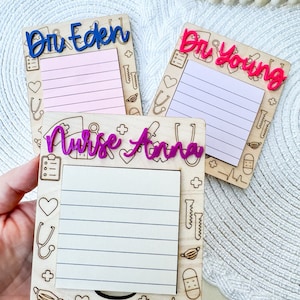 May include: Three wooden notepads with a medical theme. The notepads have a white background with black outlines of medical symbols. The top notepad has the text "Dr. Eden" in blue lettering. The middle notepad has the text "Dr. Young" in pink lettering. The bottom notepad has the text "Nurse Anna" in purple lettering.
