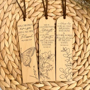 Könnte beinhalten: Drei Lesezeichen aus Holz mit braunen Lederschnüren. Jedes Lesezeichen hat eingravierten Text und botanische oder Schmetterlingsillustrationen. Der Text enthält inspirierende Zitate aus Sprüche 31:28, 31:25 und 31:31. Die Lesezeichen werden auf einer gewebten, natürlich getönten Matte präsentiert.