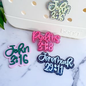 May include: A collection of decorative bag charms with religious scripture quotes. The charms feature the text "John 3:16", "Psalms 24:8", and "Jeremiah 29:11" in various colors and styles, including glitter accents.