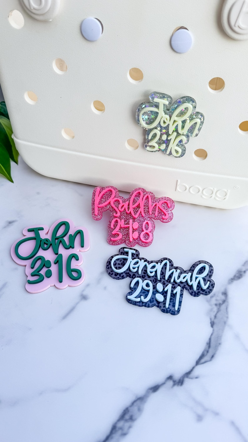 May include: A collection of decorative bag charms with religious scripture quotes. The charms feature the text "John 3:16", "Psalms 24:8", and "Jeremiah 29:11" in various colors and styles, including glitter accents.