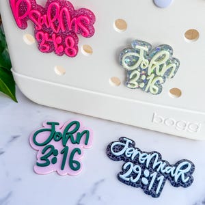 May include: Four decorative charms with religious text, including "Psalms 23:6", "John 3:16", and "Jeremiah 29:11". The charms are in various colors, including pink, green, and blue, and are embellished with glitter. They are designed to be attached to a white tote bag.