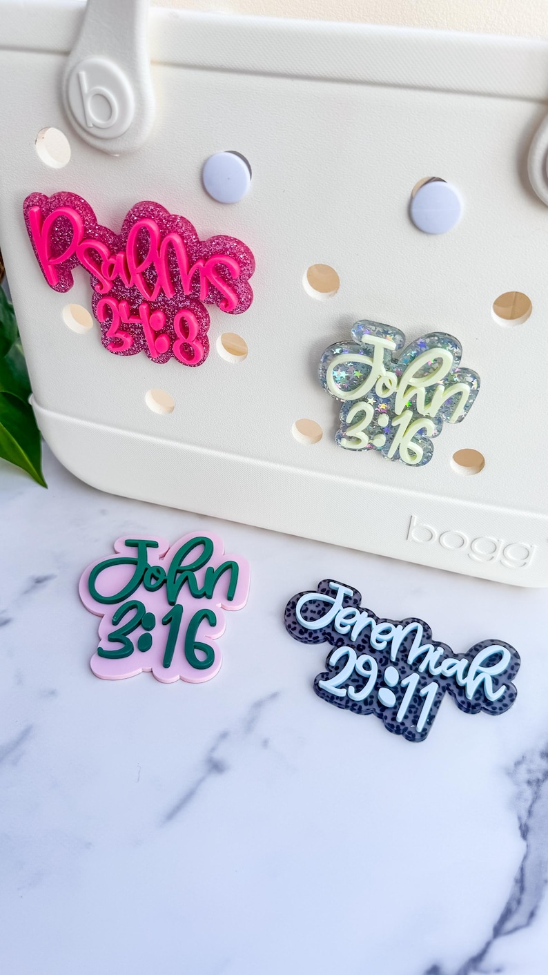 May include: Four decorative charms with religious text, including "Psalms 23:6", "John 3:16", and "Jeremiah 29:11". The charms are in various colors, including pink, green, and blue, and are embellished with glitter. They are designed to be attached to a white tote bag.