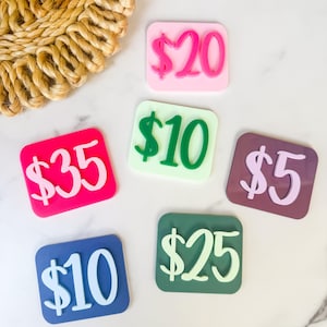 Pode incluir: Uma coleção de etiquetas de preço quadradas e coloridas. Cada etiqueta exibe um valor diferente em dólares, incluindo $5, $10, $20, $25 e $35. As etiquetas estão dispostas sobre uma superfície branca.