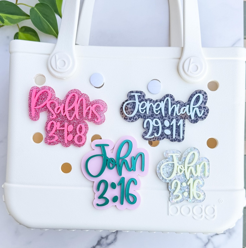 May include: A white tote bag with decorative charms. The charms feature Bible verses in various colors and designs, including pink glitter, leopard print, and pastel hues. The bag has a handle and the brand name "bogg" is visible.