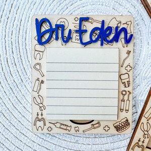 Könnte beinhalten: Notizblock aus Holz mit einem blauen Titel "Dr. Eden" und einem linierten Notizbereich. Der Notizblock ist mit Zahnarztwerkzeugen und Zähnen verziert.