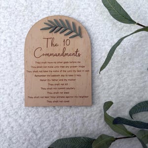 Puede incluir: Placa de madera clara con el texto "The 10 Commandments" y un diseño decorativo de hojas. El texto está en una fuente clásica. El fondo es una superficie blanca texturizada.