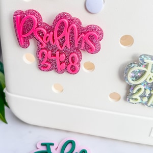 May include: Close-up of a white bag with several decorative charms. The charms feature Bible verses, including "Psalms 23:8" in pink glitter, "John 3:16" in green, and others. The bag has a circular button and several holes.