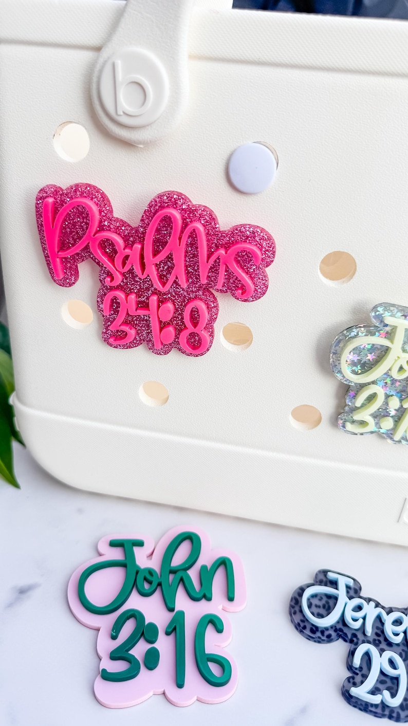 May include: Close-up of a white bag with several decorative charms. The charms feature Bible verses, including "Psalms 23:8" in pink glitter, "John 3:16" in green, and others. The bag has a circular button and several holes.