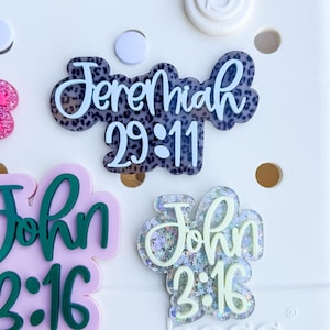 May include: White tote bag with several decorative acrylic charms. The charms include the text "Jeremiah 29:11" in a black and white leopard print design, and "John 3:16" in green and glittery designs. The bag has a white background.