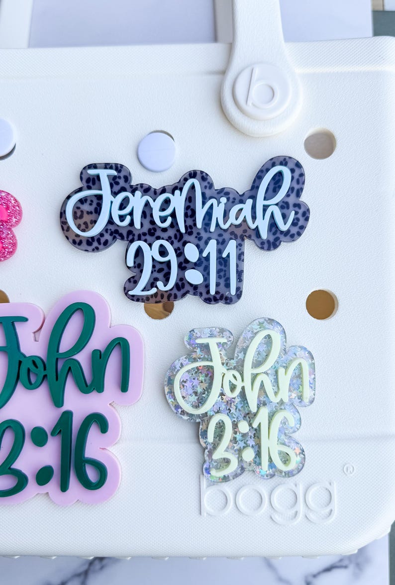 May include: White tote bag with several decorative acrylic charms. The charms include the text "Jeremiah 29:11" in a black and white leopard print design, and "John 3:16" in green and glittery designs. The bag has a white background.