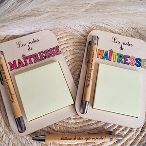 Peut inclure: Deux blocs-notes en bois avec le texte "Les notes de MAÎTRESSE" en lettres colorées. Chaque bloc-notes contient un bloc de notes adhésives jaunes et un stylo en bois. Les stylos ont des détails argentés et du texte. Les blocs-notes sont sur une surface tissée.