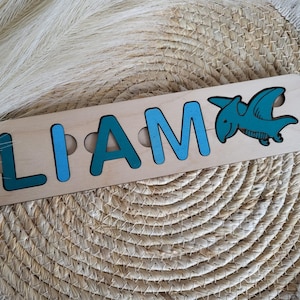 Peut inclure: Un puzzle à lettres en bois rectangulaire affichant le prénom LIAM avec des lettres bleu clair, où la dernière lettre est remplacée par une figurine de ptérodactyle bleu sarcelle. Les pièces sont insérées dans un socle en bois clair naturel.