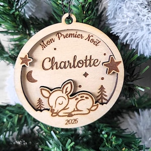 Peut inclure: Ornement de Noël en bois avec l'inscription "Mon Premier Noël" et le prénom "Charlotte". L'ornement représente un cerf, des étoiles et un croissant de lune. L'année 2025 est également incluse. L'ornement est suspendu à un sapin de Noël.