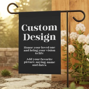 May include: A black garden flag with the words "Custom Design" in white. The flag is displayed on a black metal stand in a garden setting. The flag also includes the text "Honor your loved one and bring your vision to life. Add your favorite picture, saying, name and dates."
