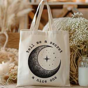 May include: A white canvas tote bag with black text that reads "tell me a secret a nice one" and a black crescent moon with stars inside.