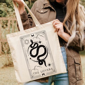 May include: A beige tote bag with a black and white illustration of a snake and the words "The Lovers" on the front.