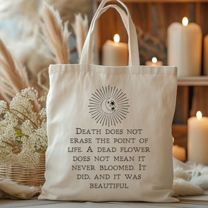 May include: A white canvas tote bag with black text that reads "Death does not erase the point of life. A dead flower does not mean it never bloomed. It did, and it was beautiful." The bag features a black and white illustration of a crescent moon with stars and sun rays.
