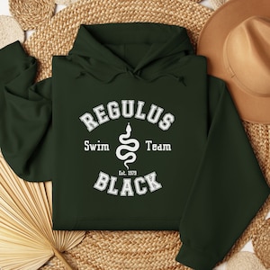 以下が含まれることがあります： 「Regulus Swim Team Est. 1979 Black」と蛇のデザインが書かれた白いグラフィックプリントが施された濃い緑色のフード付きスウェットシャツです。