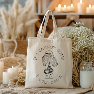 May include: A white canvas tote bag with black text and a graphic of a tree with a snake wrapped around it. The text reads "If I cannot climb I will grow".