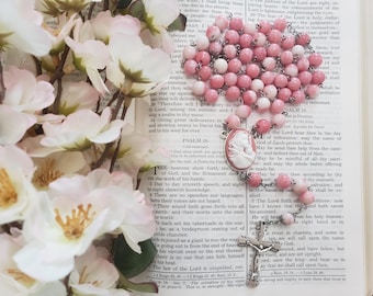 May include: A rosary featuring round pink and white beads, a silver-toned metal crucifix, and a central oval cameo pendant depicting a profile portrait. The rosary is arranged on an open book with printed text, accompanied by white flowers.