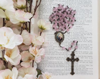 May include: A rosary featuring light pink beads and a dark metal chain. The centerpiece is an oval medallion depicting a religious figure, and the crucifix is made of dark metal. The rosary is placed over an open book with printed text.
