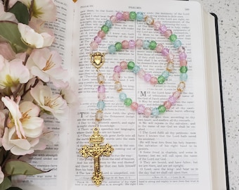 May include: A rosary featuring translucent pastel pink, green, and blue beads arranged in a circular pattern. The rosary includes a gold-toned metal centerpiece depicting a heart and a detailed gold-toned crucifix with ornate filigree edges.