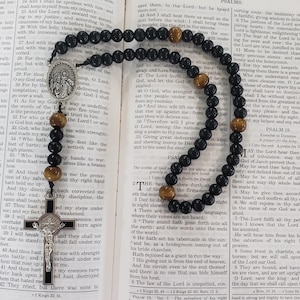 May include: A black beaded rosary with brown accent beads and a silver crucifix lies open on a book. The rosary features a silver centerpiece with a religious figure. The book's pages are filled with text.