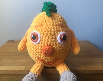 May include: A crocheted toy in the shape of a smiling, orange creature with large eyes and a green tuft on top. The toy has small arms and legs, and is sitting on a wooden surface.
