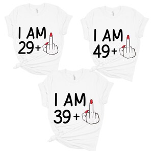 May include: Three white t-shirts with black text and a red middle finger graphic. The text on the shirts reads "I am 29+", "I am 39+", and "I am 49+".