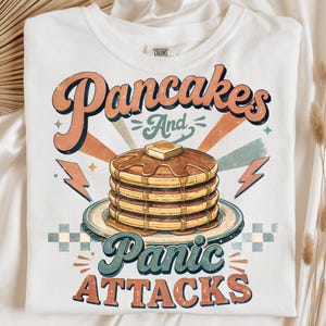 May include: A white t-shirt featuring a graphic print of a stack of pancakes with syrup and a pat of butter on a plate. The text reads "Pancakes And Panic ATTACKS" in a retro font, surrounded by lightning bolts, stars, and checkered patterns.