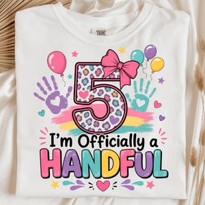 Könnte beinhalten: Ein weißes T-Shirt mit einer großen Zahl 5 in einem bunten Leopardenmuster, gekrönt von einer rosa Schleife. Das Design enthält violette und türkisfarbene Handabdrücke, Luftballons, Herzen, Sterne und den Text I'm Officially a HANDFUL.