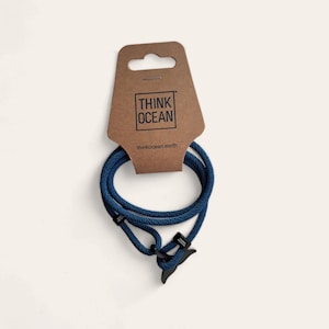 Pode incluir: Um bracelete de corda azul escuro com um amuleto de cauda de baleia. O bracelete está embalado em um cartão marrom com o texto "Think Ocean" e "thinkocean.earth".