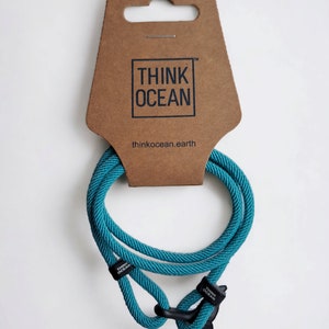 Pode incluir: Pulseira de corda azul turquesa com um fecho de cauda de baleia e as palavras "Think Ocean" no fecho. A pulseira é exibida em um cabide de cartão castanho com as palavras "Think Ocean" e "thinkocean.earth" impressas nele.