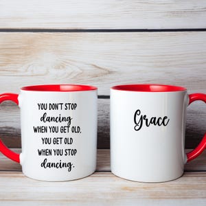 May include: Two white ceramic mugs with red interiors and handles. One mug has the quote "YOU DON'T STOP dancing WHEN YOU GET OLD, YOU GET OLD WHEN YOU STOP dancing." The other mug says "Grace."