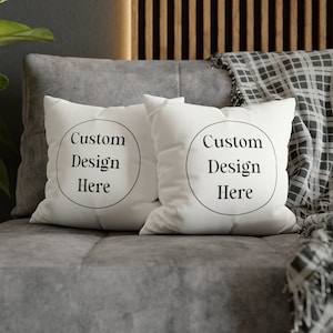 May include: Two square white throw pillows featuring a black circular outline and the black text Custom Design Here printed in the center of each pillow. The pillows are positioned side by side, displaying a clean, plain fabric surface.