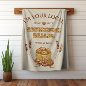 May include: A beige kitchen towel with the text "I'm your local home made sourdough dealer wake & bake" in brown lettering. The towel features a basket of bread and wheat stalks.