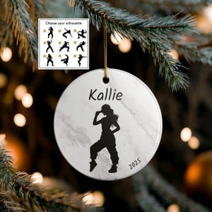 May include: A white marble-patterned ceramic ornament with a black silhouette of a dancer. The ornament has the name "Kallie" and the year "2025" printed on it. A selection of dance silhouettes is shown in a small box. The ornament hangs from a gold string.