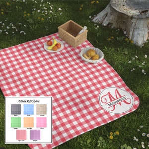 May include: A red and white checkered picnic blanket with a personalized monogram. The blanket is spread on green grass, with a picnic basket, fruit, and baked goods. Color options are displayed in a small box.